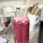 straight-on-view-of-large-mason-jar-full-of-blueberry-lemonade-on-ktichen-counter-with-wood-spoon-sticking-out